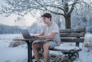 kluk který sedí v kraťasech na lavičce, má notebook ale venku je zima sníh a mráz je spokojený, v pozadí je zasněžený sad